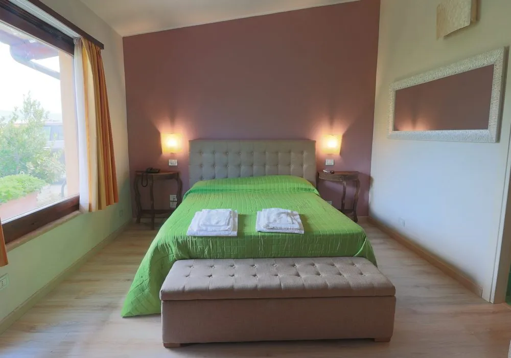 Camera con copriletto verde, panca capitonné ai piedi del letto e finestra affacciata sulla valle