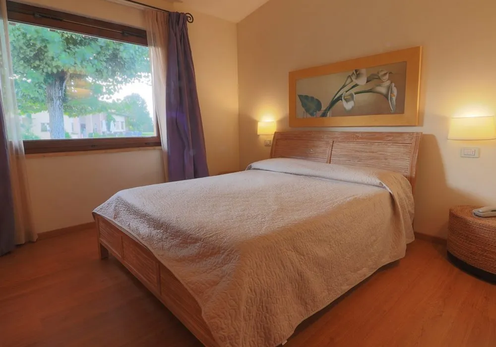 Camera con testata in midollino, quadro floreale, tende viola e finestra sul giardino