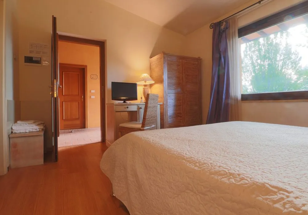 Camera con armadio e scrittoio in midollino, porta aperta sul corridoio e letto con copriletto bianco