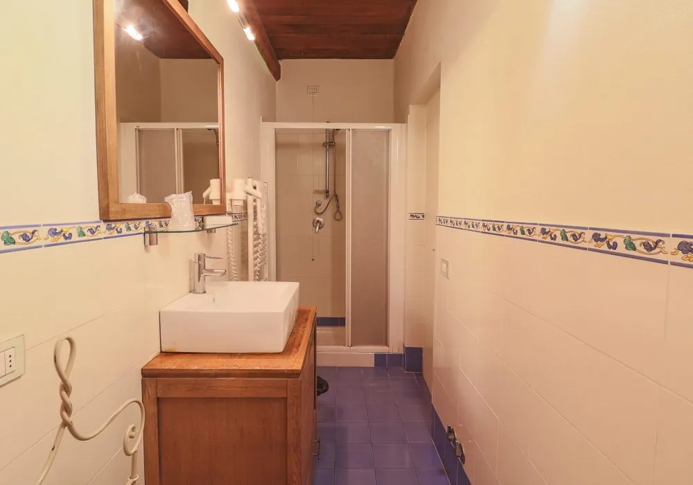 Bagno con pavimento e fascia a maiolica blu, soffitto a travi in legno e box doccia in vetro