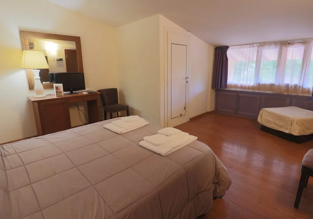Camera con letto e asciugamani piegati, scrittoio con TV e specchio, secondo letto sullo sfondo