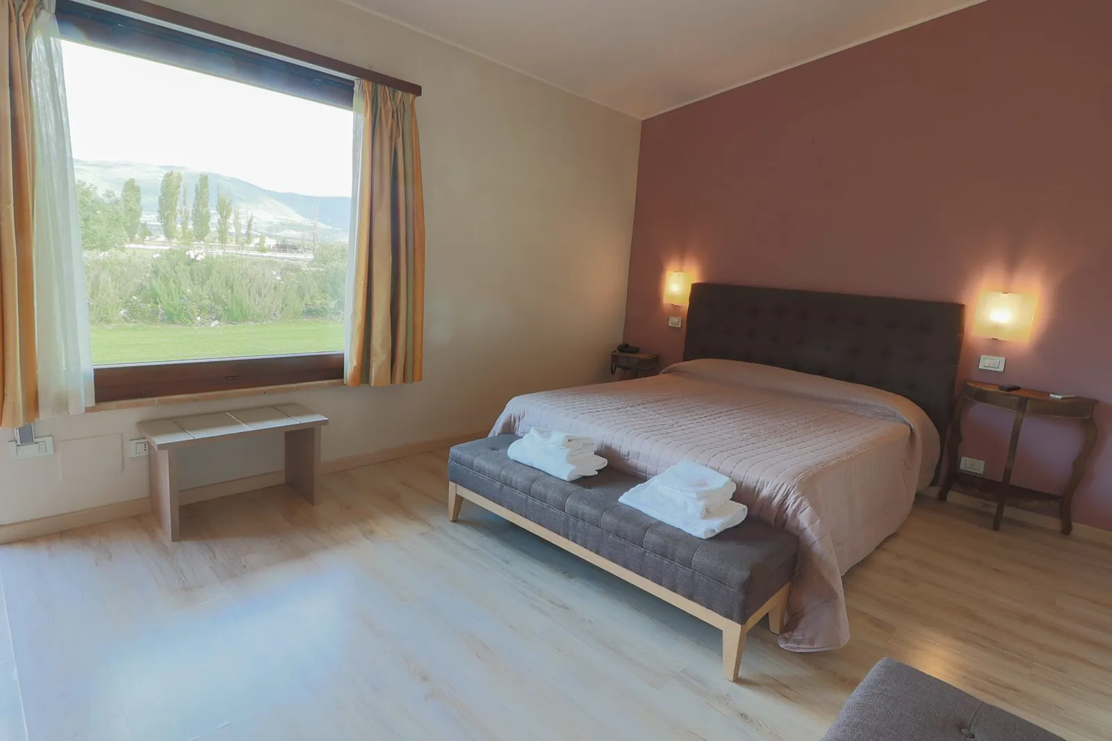 Room with a large window onto the park and hills, dusty-pink wall, upholstered headboard and a bench at the foot of the bed