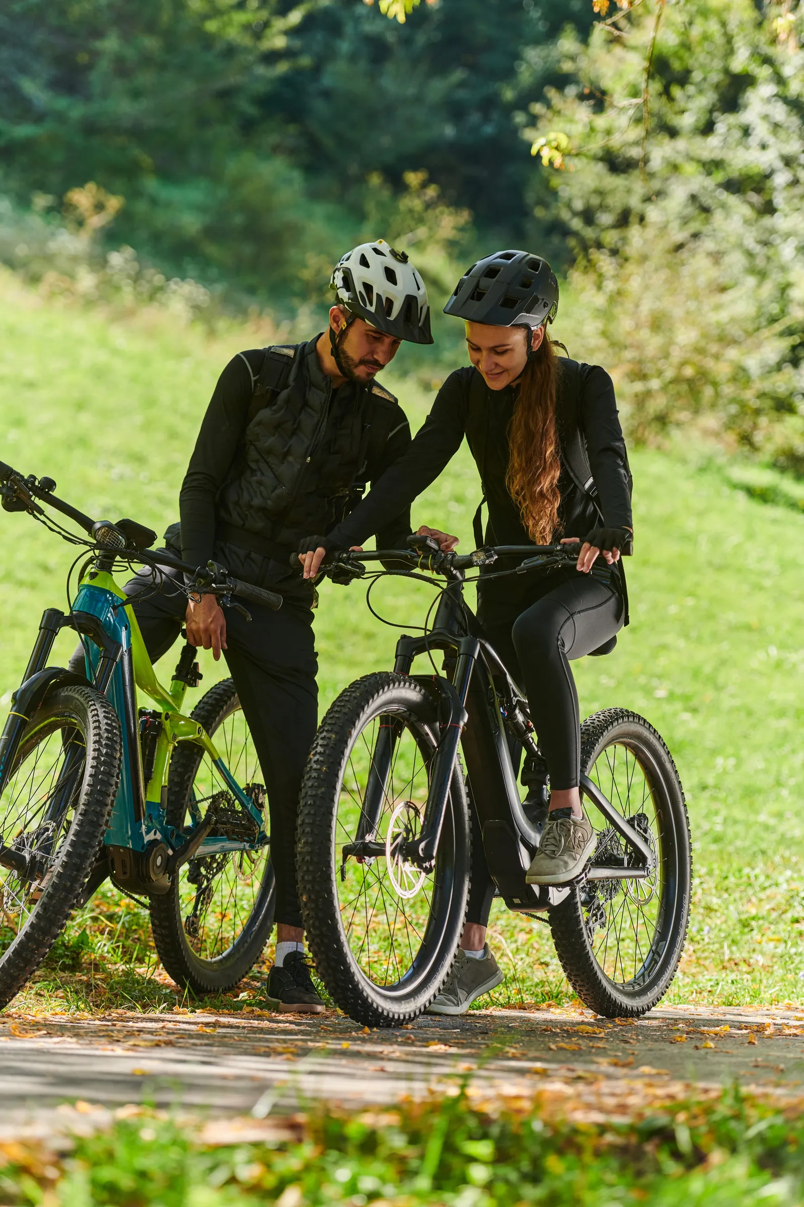 Two cyclists with helmets pause to check a bike display, on a path through the park