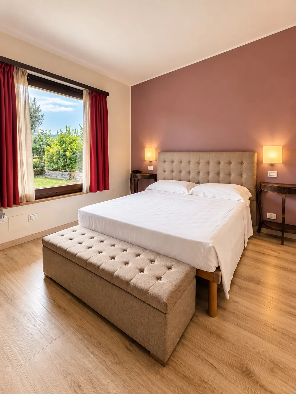 Room with a dusty-pink accent wall behind a tufted upholstered headboard, white bedding and red curtains at the window