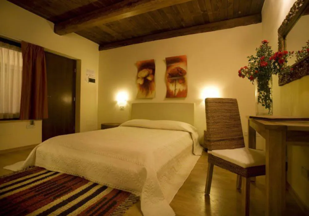 Camera con soffitto a travi, letto con copriletto bianco, due quadri floreali, sedia in midollino e specchio con vaso di rose