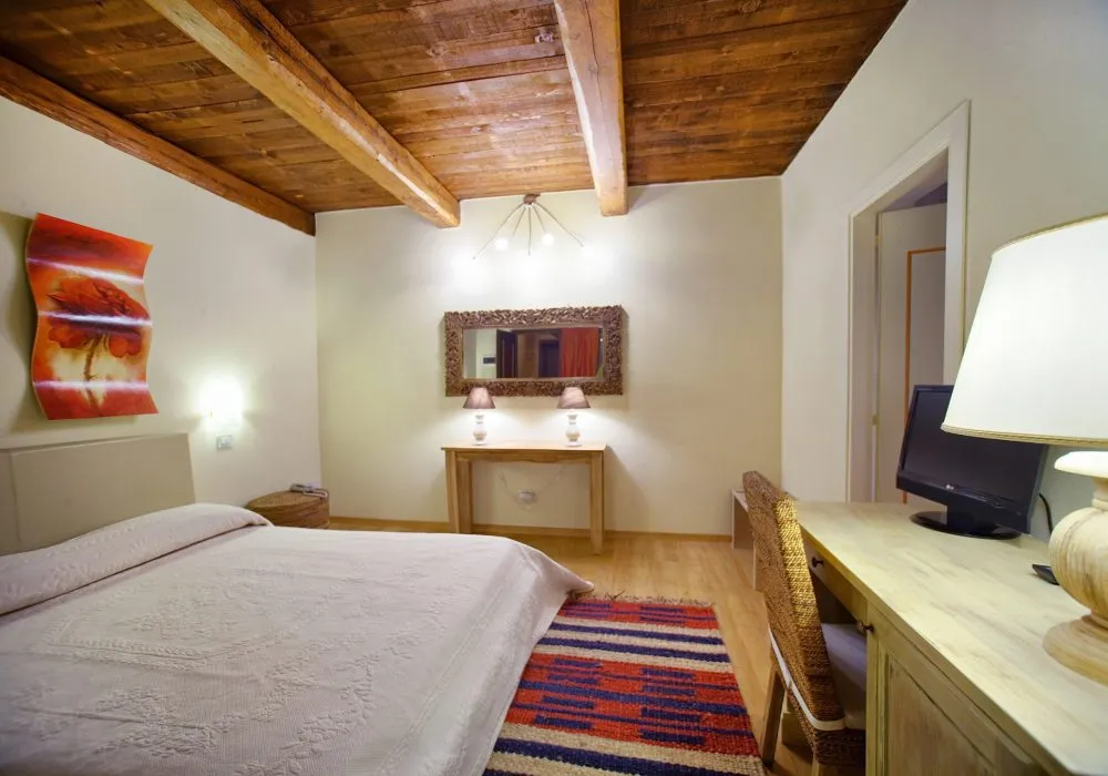 Camera con soffitto a travi a vista, testata beige, console con specchio e due lampade, scrivania con monitor e tappeto a righe