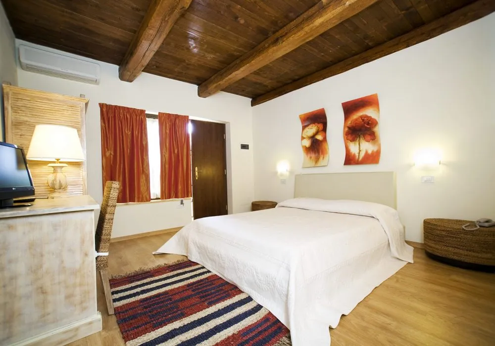 Camera con soffitto a travi, tende rosse, letto con copriletto bianco, due quadri floreali e climatizzatore a parete