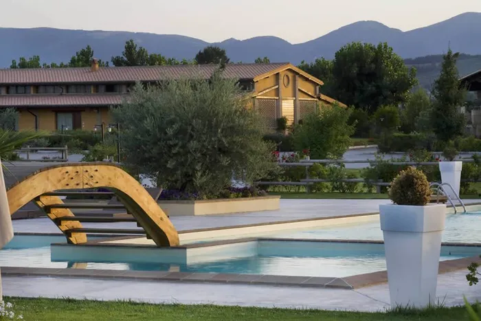 Outdoor pool at dusk with a wooden footbridge, olive tree and hills behind