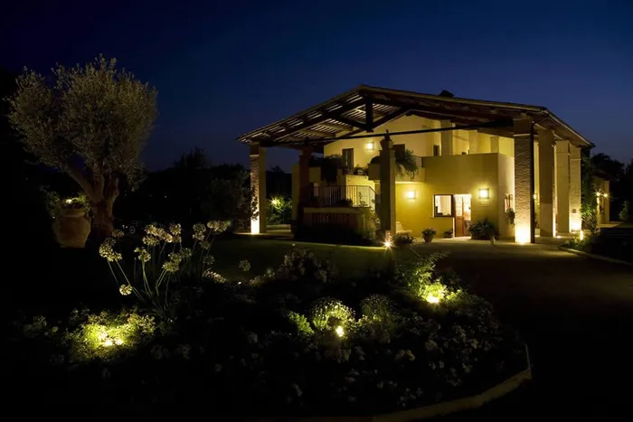 Ingresso illuminato di notte, con un ulivo e un'aiuola fiorita in primo piano