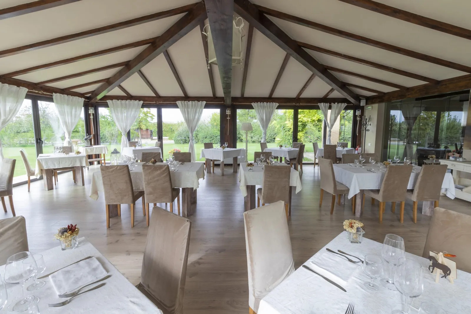 Sala ristorante con travi a vista, tavoli apparecchiati in bianco e vetrate sul giardino