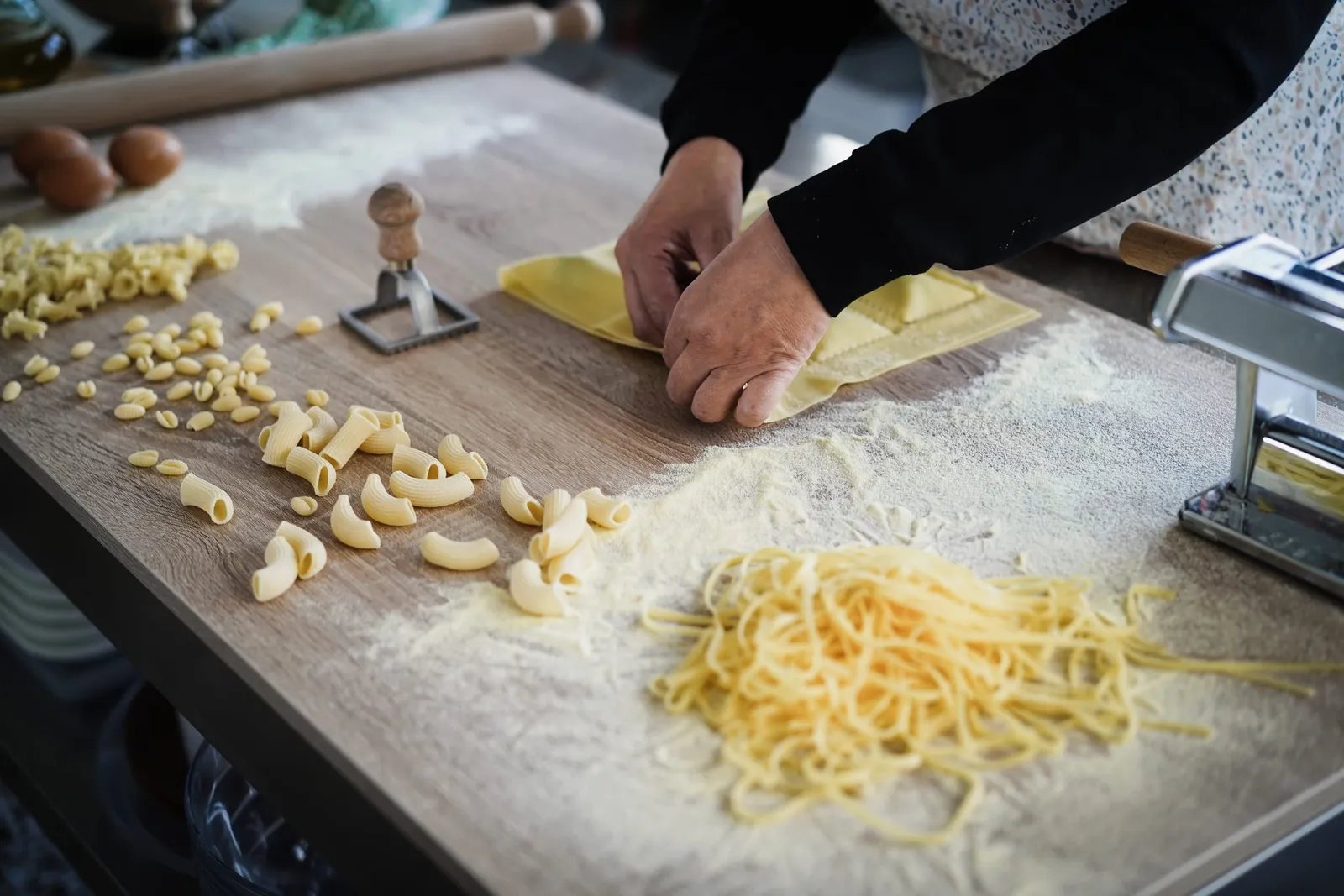 Hands working fresh homemade pasta on a wooden board, among tagliatelle, eggs and a small pasta press