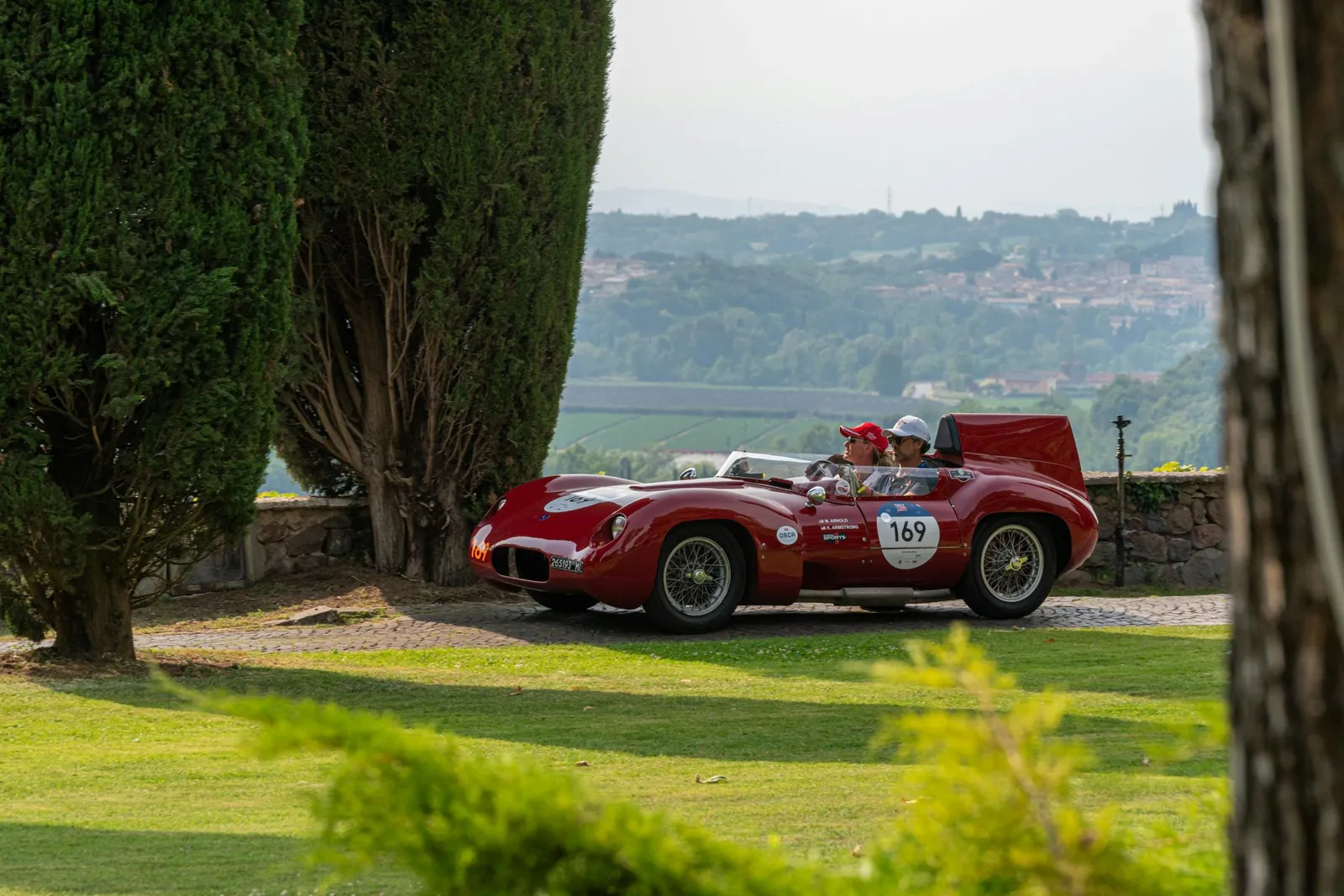 Auto d'epoca rossa da competizione con due persone a bordo, su un prato fra cipressi e colline