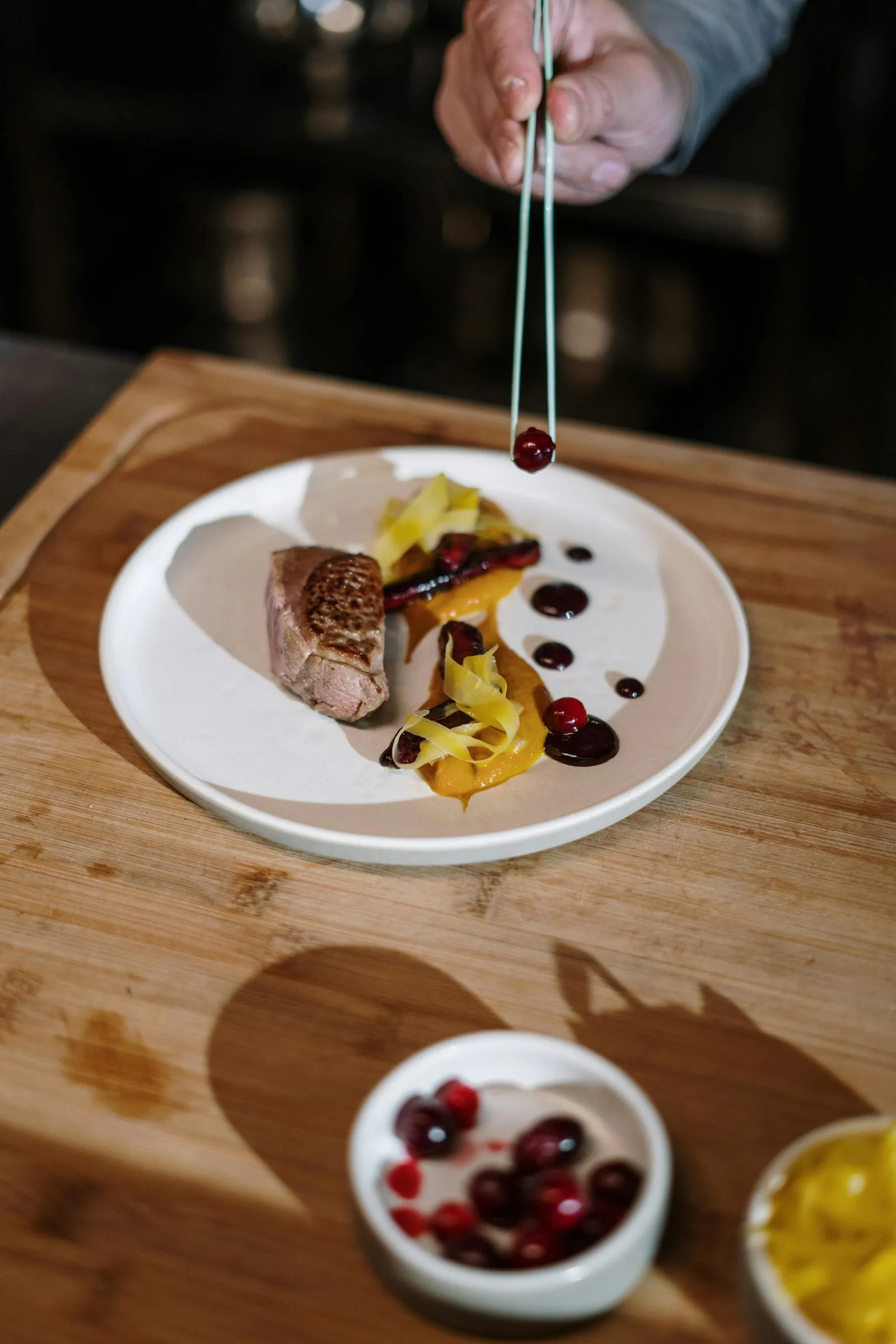 A chef's hand placing a cranberry with tweezers on a plate of meat