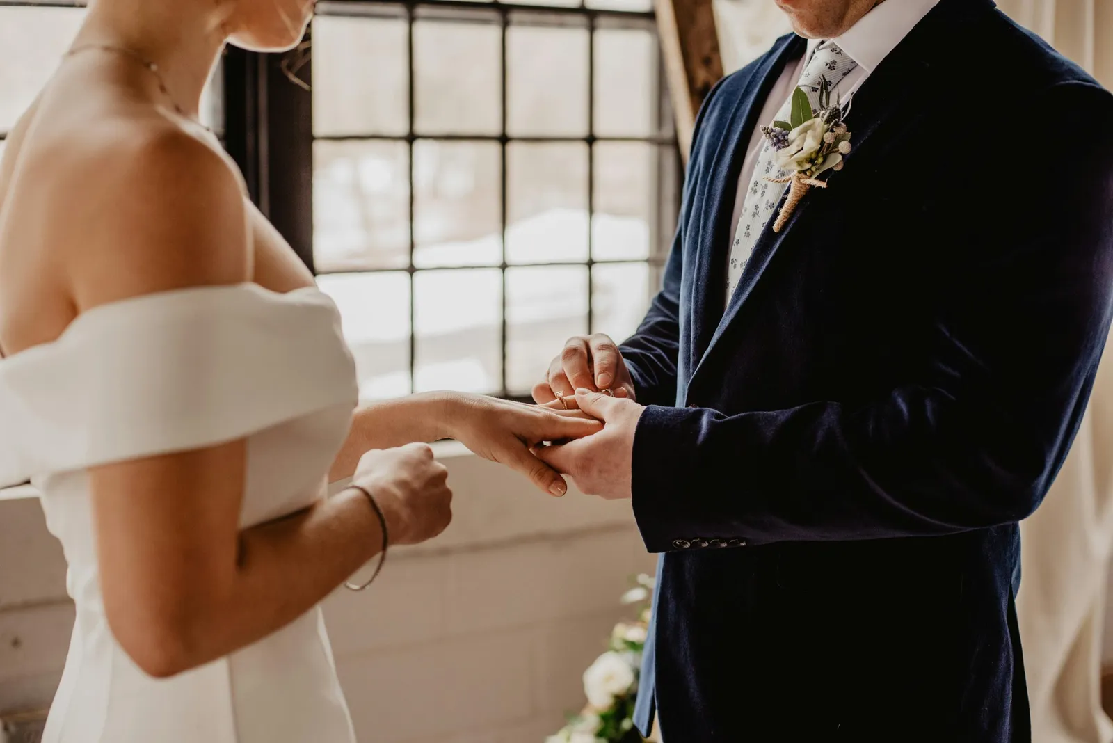 Bride and groom exchanging rings by a tall window, groom in navy jacket