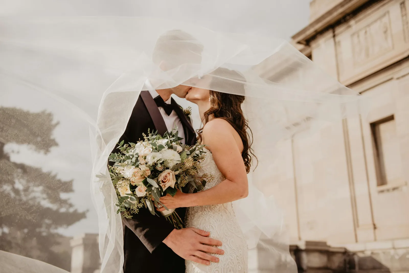Bride and groom kissing under a windblown veil, holding a pale rose bouquet