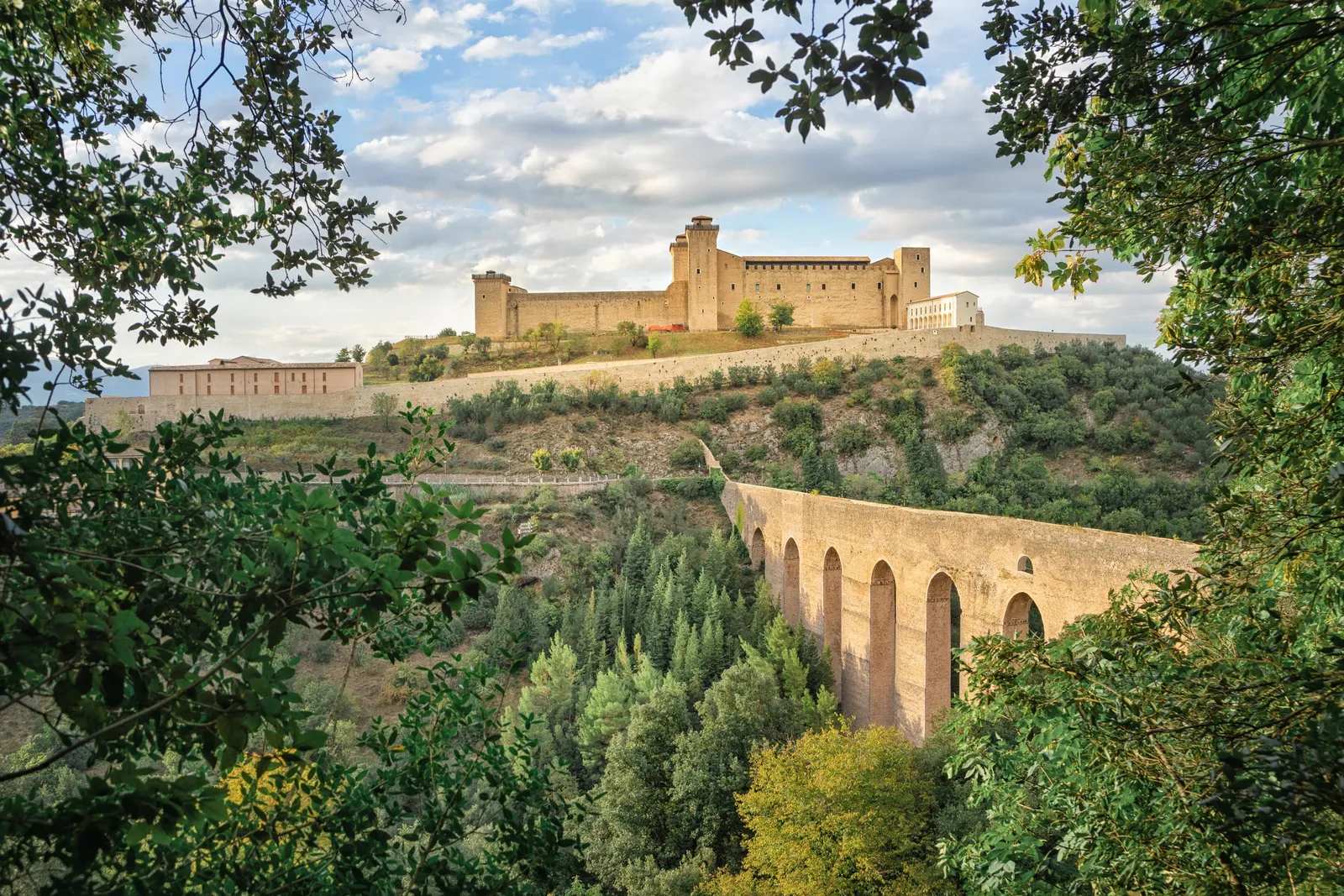The Rocca Albornoziana fortress of Spoleto on the hilltop, with the Ponte delle Torri bridge and woods in the foreground
