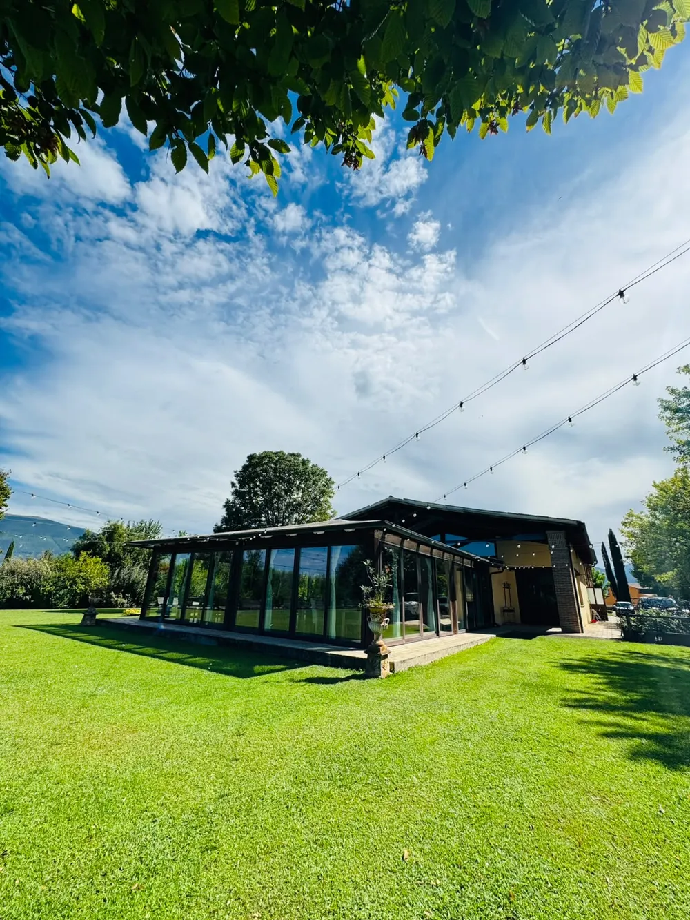 Glass-walled veranda with dark wood beams and rows of hanging lights, green lawn and hills beyond